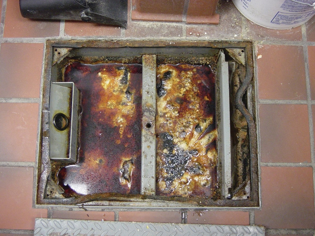  A picture of the floor with a grease trap open in the middle of the floor. The grease trap is a square hole in the floor. This square floor is filled with solidified grease.
